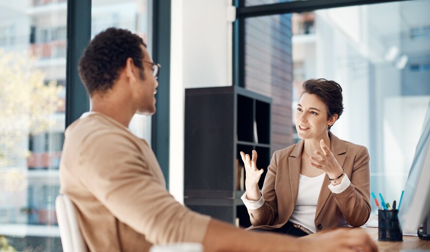 Eine Frau und ein Mann sitzen sich in einem modernen Büro gegenüber und führen ein engagiertes, gestenreiches Gespräch.